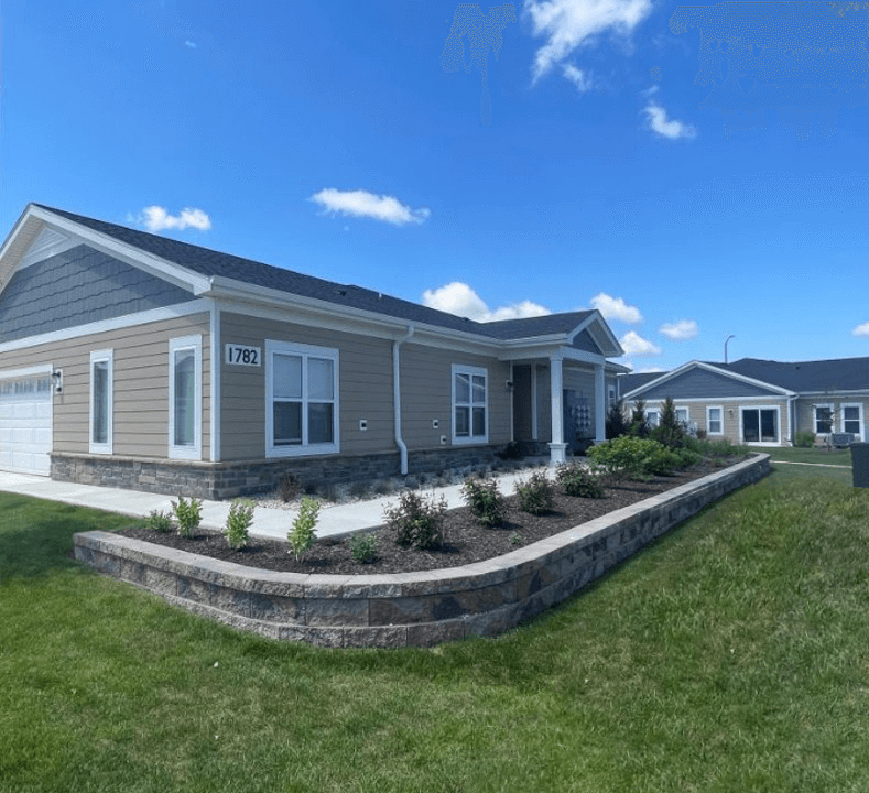 Single-story beige house with white trim and stone accents, large windows, and a well-maintained garden bed in front. The house number 1782 is visible. The yard has green grass and a paved walkway under a clear blue sky.