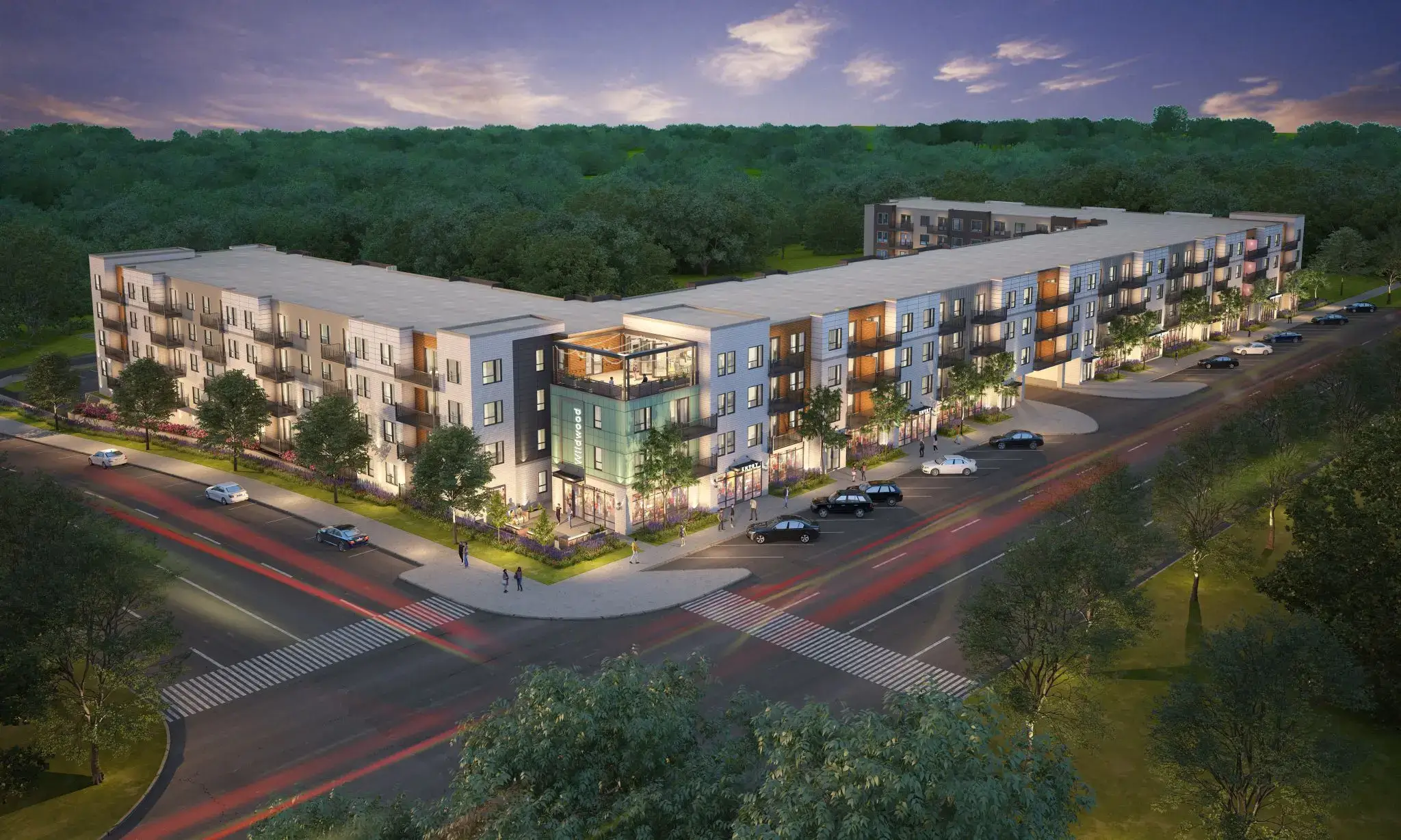 A modern four-story apartment complex with well-lit windows at dusk, surrounded by trees and roads with streaks of car lights, viewed from above at a corner intersection.