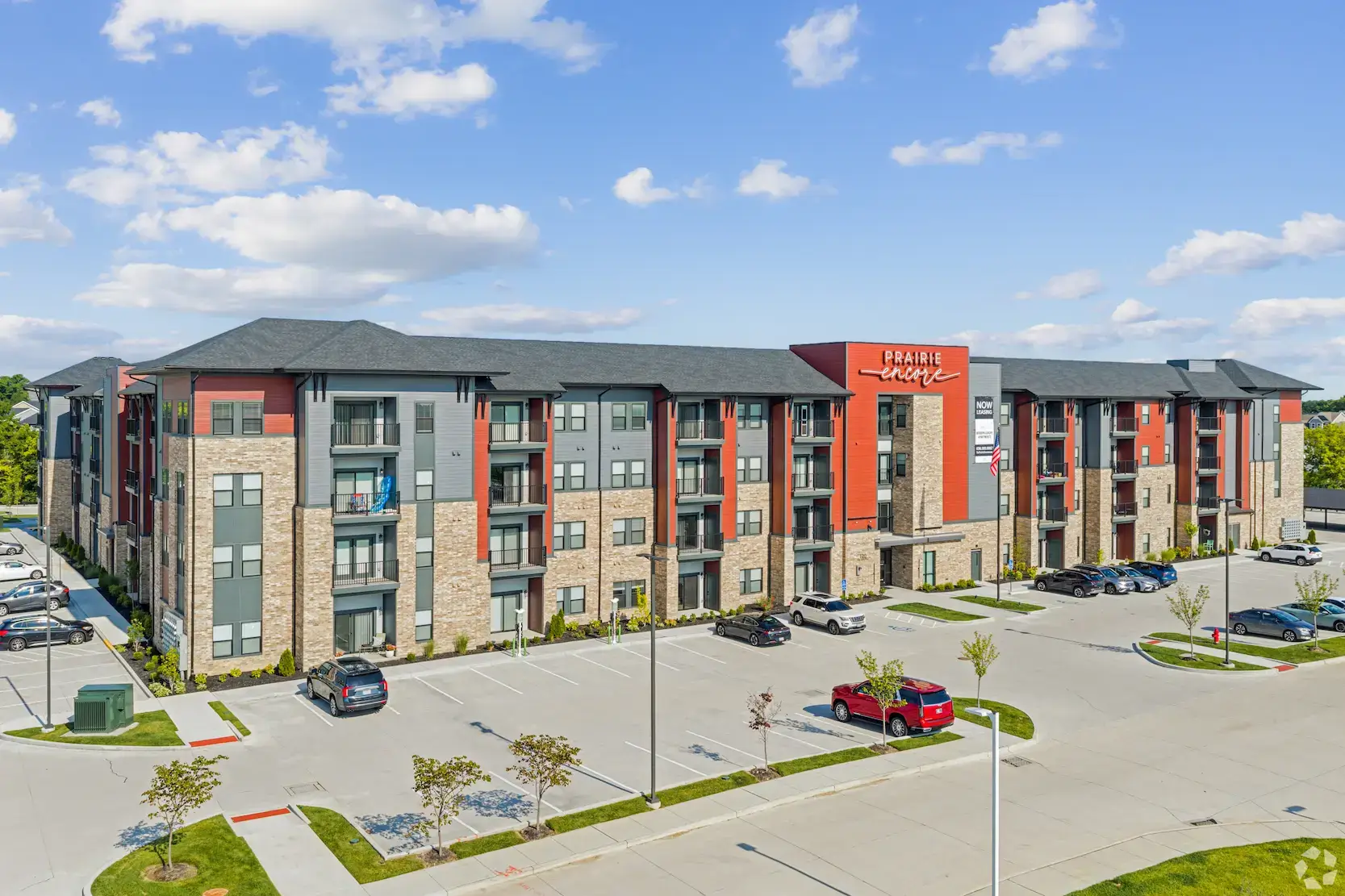 A modern four-story apartment building with balconies, stone and red siding, labeled Prairie Village. Several parked cars and small trees are in the parking lot under a blue sky with scattered clouds.